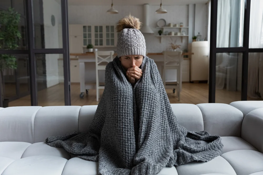 Woman bundled in a thick blanket and winter hat, sitting on a couch in a cold home waiting for furnace repair in King and Snohomish Counties.