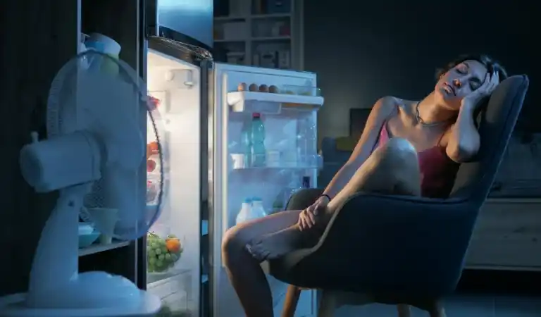 Woman sitting in front of an open refrigerator at night trying to cool down due to a broken air conditioner in her home.